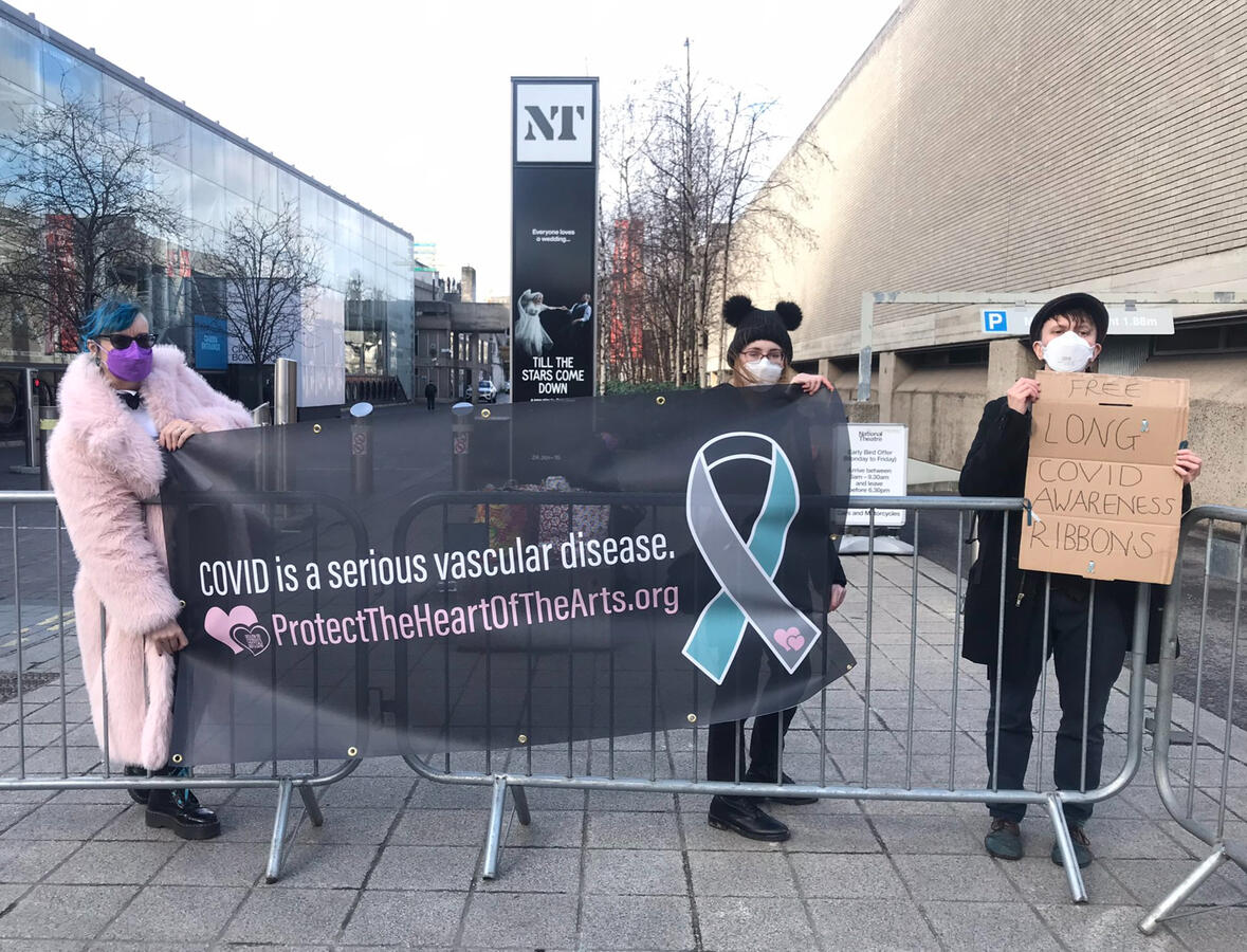 Protect the Heart of the Arts banner Three members of Protect the Heart of the Arts wearing respirators, holding up a banner that reads "Covid is a serious vascular disease" with the Long Covid awareness ribbon and link/QR code to protecttheheartofthearts.org