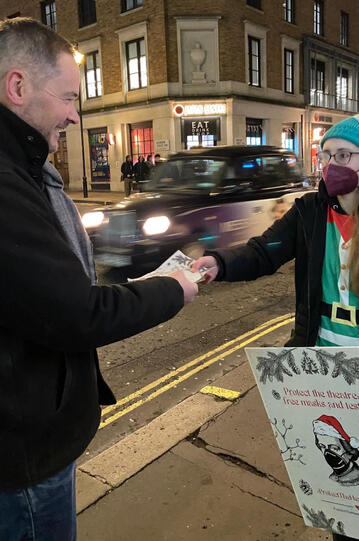 A masked person in a holiday costume and a masks and holding up a sign and offering a mask to a person on the street
